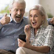 Excited surprised retired couple holding mobile phone, staring at gadget screen, shouting for joy with winner hands, celebrating victory, success, win, enjoying leisure, entertainment at home